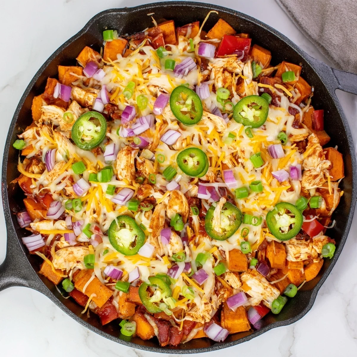 Sizzling skillet dinner with BBQ chicken, sweet potatoes, and a rich blend of cheddar and mozzarella, topped with fresh cilantro and jalapeños for a spicy kick. Sweet Potato Skillet with BBQ Chicken & Cheese.